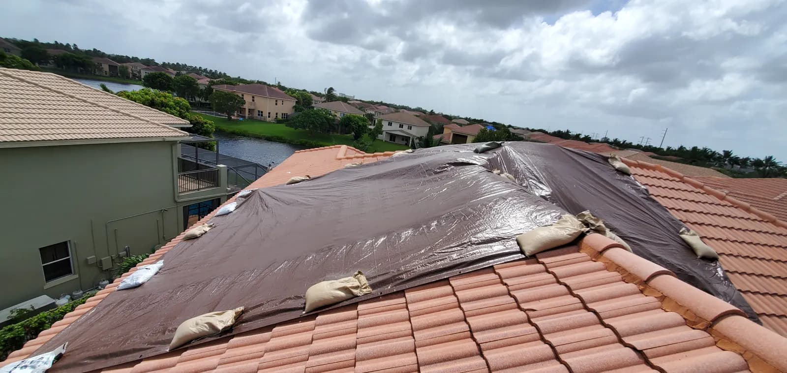 Emergency tarp and sandbags on a tile roof in a townhome community, with two-story buildings and a lake beyond