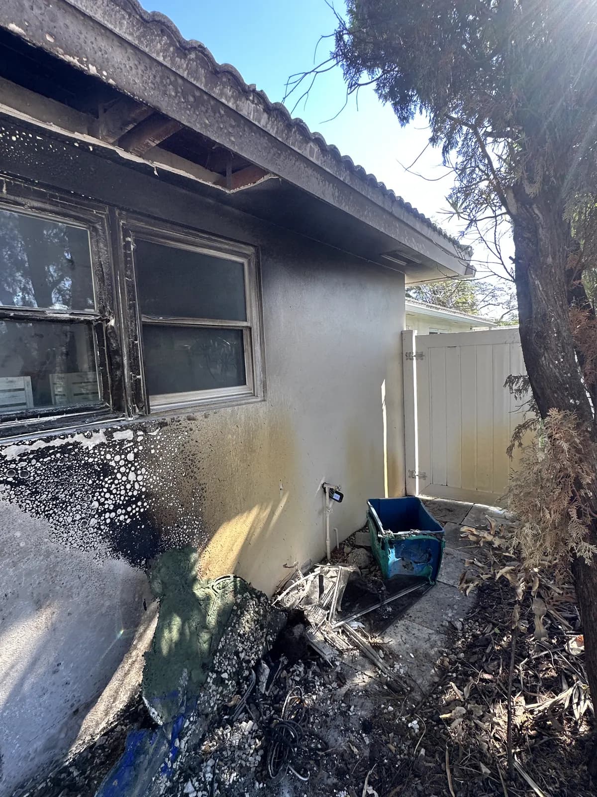 Fire-damaged home exterior with scorched stucco, burned eaves and charred debris beneath the windows