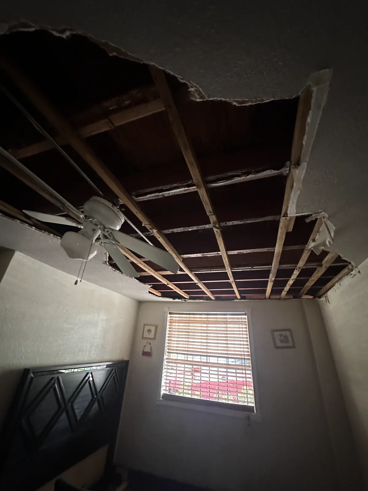 Bedroom ceiling collapsed from water damage, exposing the wood joists above a ceiling fan and window