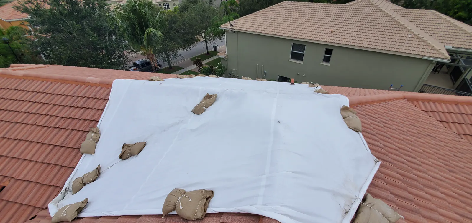 White tarp weighted with sandbags on a terracotta tile roof next to a two-story multi-unit building